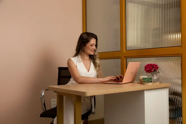O consultório com a mesa de madeira, o notebook, flores e a janela de vidro canelado